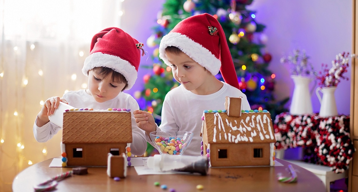 two young boys with santa hats on each building a gingerbread house with lighted Christmas trees in background