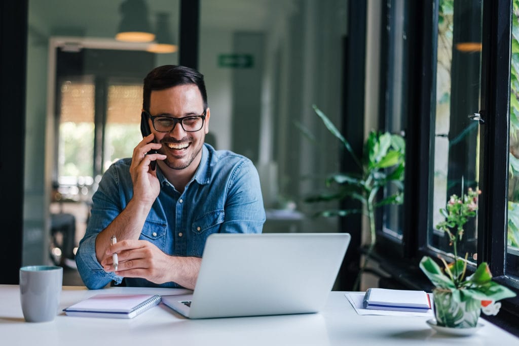 smiling young man at desk talking on phone and using laptop