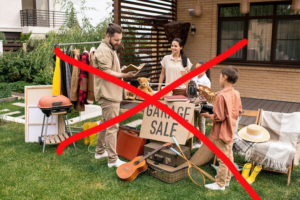 Young man with beard looking through book while choosing it at yard sale, woman and her kids waiting for customers answer. Image has a large red X across the image to indicate no more yard or garage sales.