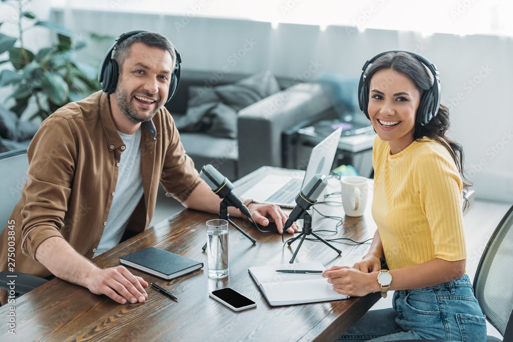 man and woman with headphones on at desk with mics in front of each diving a podcast