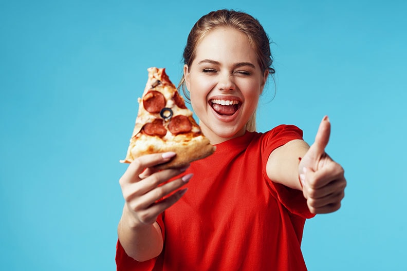 pretty woman with pizza in hands fast food eating fun. She has a red polo shirt on and a baby blue background and she's is giving the thumb up gesture, smiling. Has long ish acrylic fingernaols.