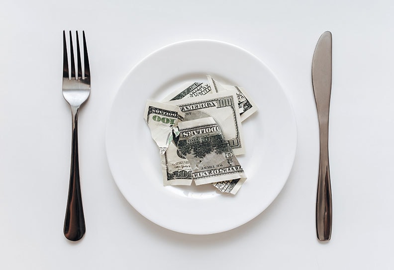 A torn old hundred-dollar bill lies on a ceramic plate between a fork and a knife on a with tablecloth. The concept of business lunch, banking, life rentier and financial well-being.