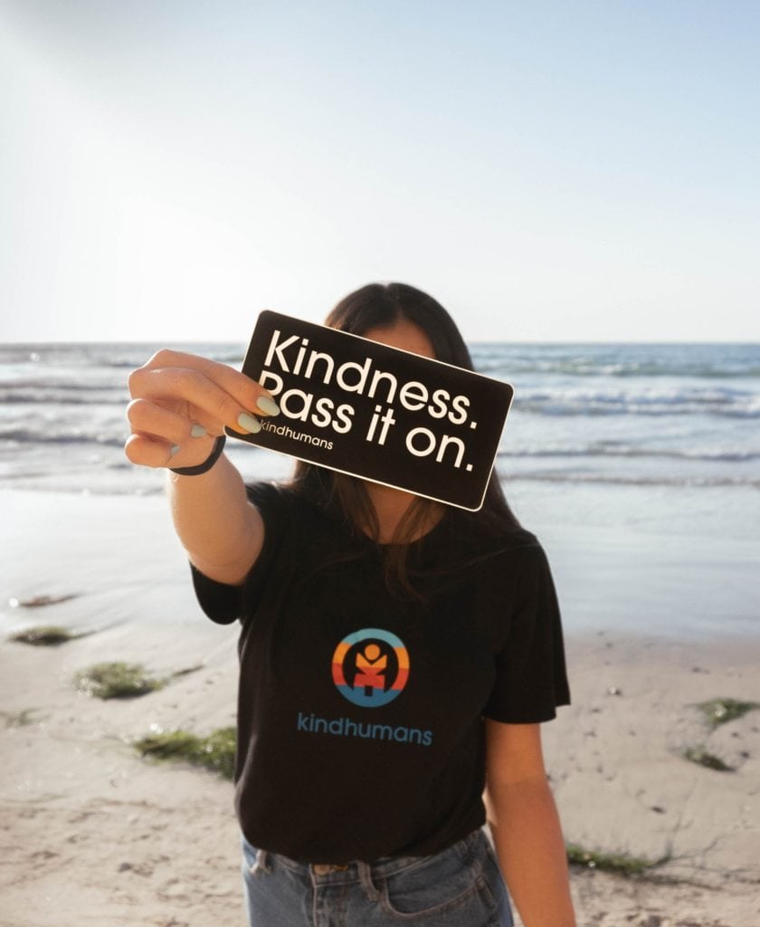 woman on a beach holding a sticker that says "kindness, pass it on" 